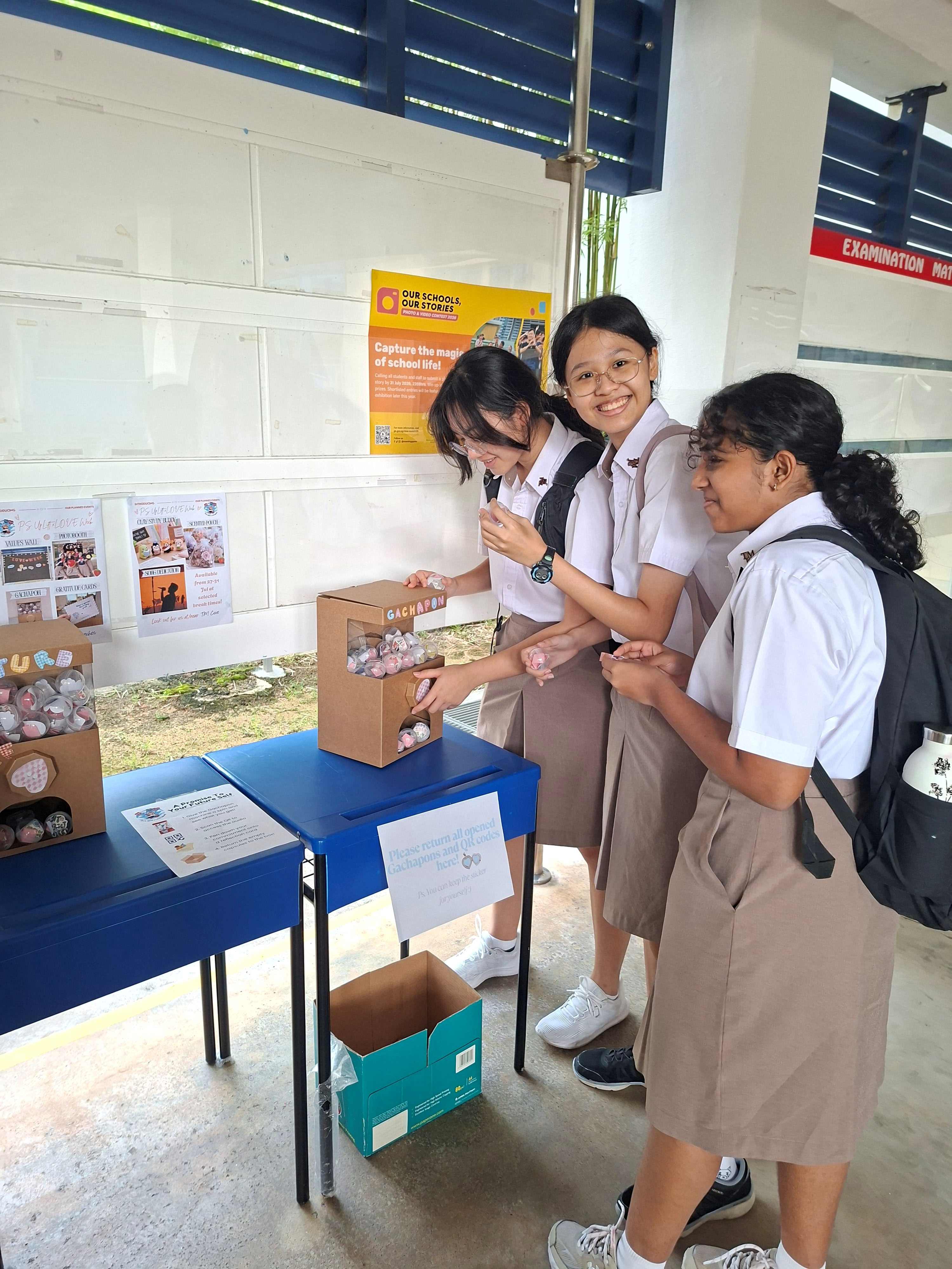 Three students in white school uniforms lean over a box labelled "Gacha" containing small capsules, smiling as they pick one up for a reflection activity.